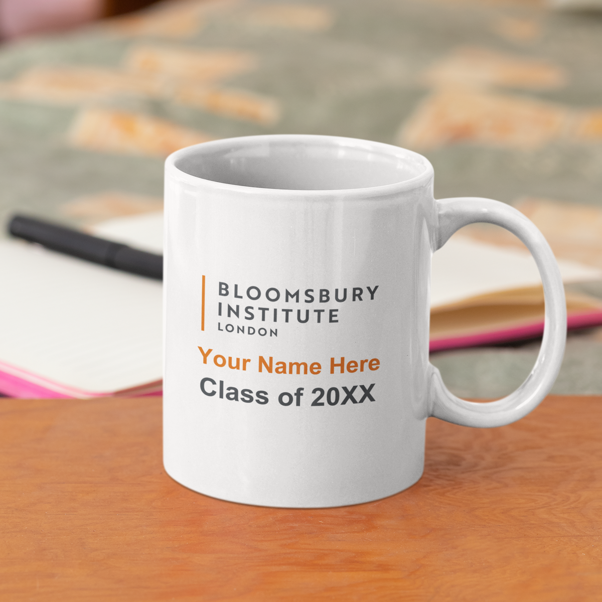 A white ceramic mug with the Bloomsbury Institute London logo and customizable text 'Your Name Here Class of 20XX' in black and orange, sitting on a wooden table with a blurred study background.