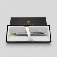 A silver and gold Cross pen presented in an open black gift box with a white insert and ribbon, photographed on a light gray background.